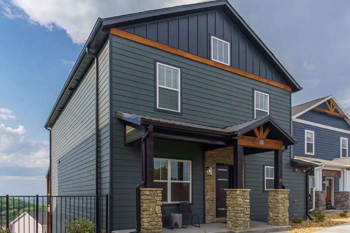 A modern exterior scene highlighting the building's striking architecture, with stone pillars, expansive windows, and a generous roof overhang. Guests are seen relaxing on the porch, surrounded by inviting outdoor seating and stunning natural views.