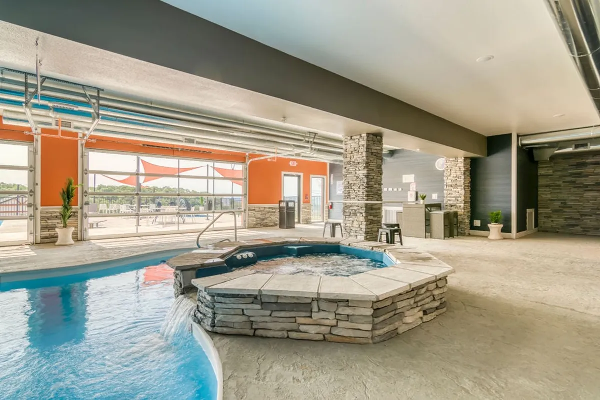A modern indoor pool area featuring a swimming pool, jacuzzi, large windows letting in natural light, a stone wall, and bar stools for casual socializing. The view outside enhances the indoor-outdoor connection.