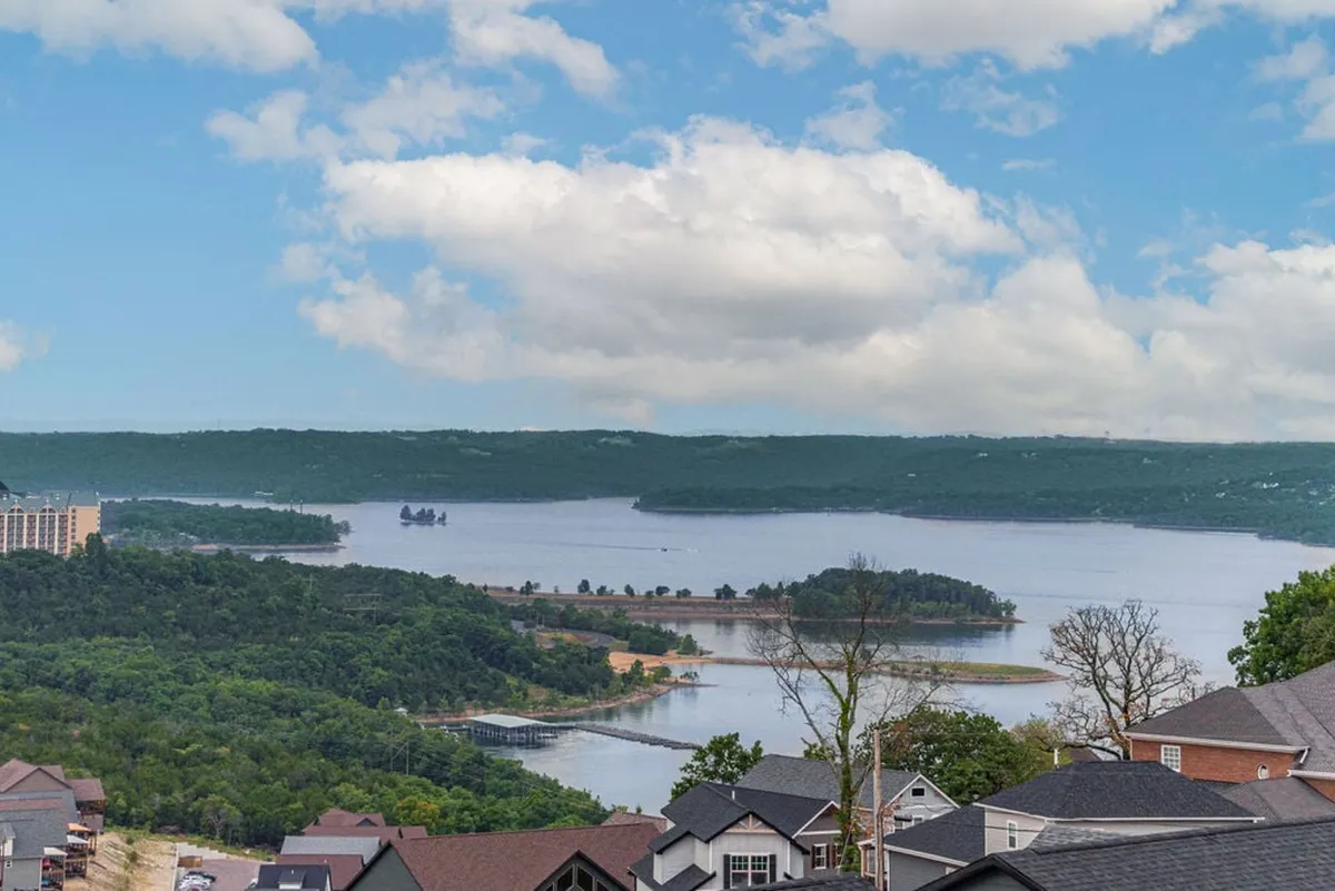 The image showcases a breathtaking, panoramic view of Table Rock Lake surrounded by lush green hills under a serene, cloudy sky. Homes dot the hillside, hinting at a peaceful lakeside community, while the tranquil water invites boating, fishing, and relaxation. The overall mood is calm and welcoming, perfect for unwinding and connecting with nature.