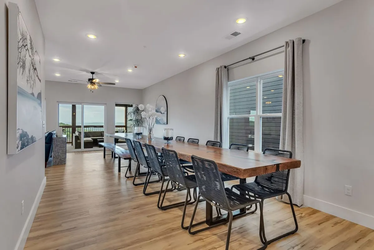This image features a spacious dining area with a large wooden dining table, comfortable seating, and large windows that allow natural light to flood the room. Decorative artwork adds a stylish touch, while the view from the windows hints at a scenic outdoor setting. The table is set up to accommodate a large group, making it ideal for family gatherings or group celebrations.