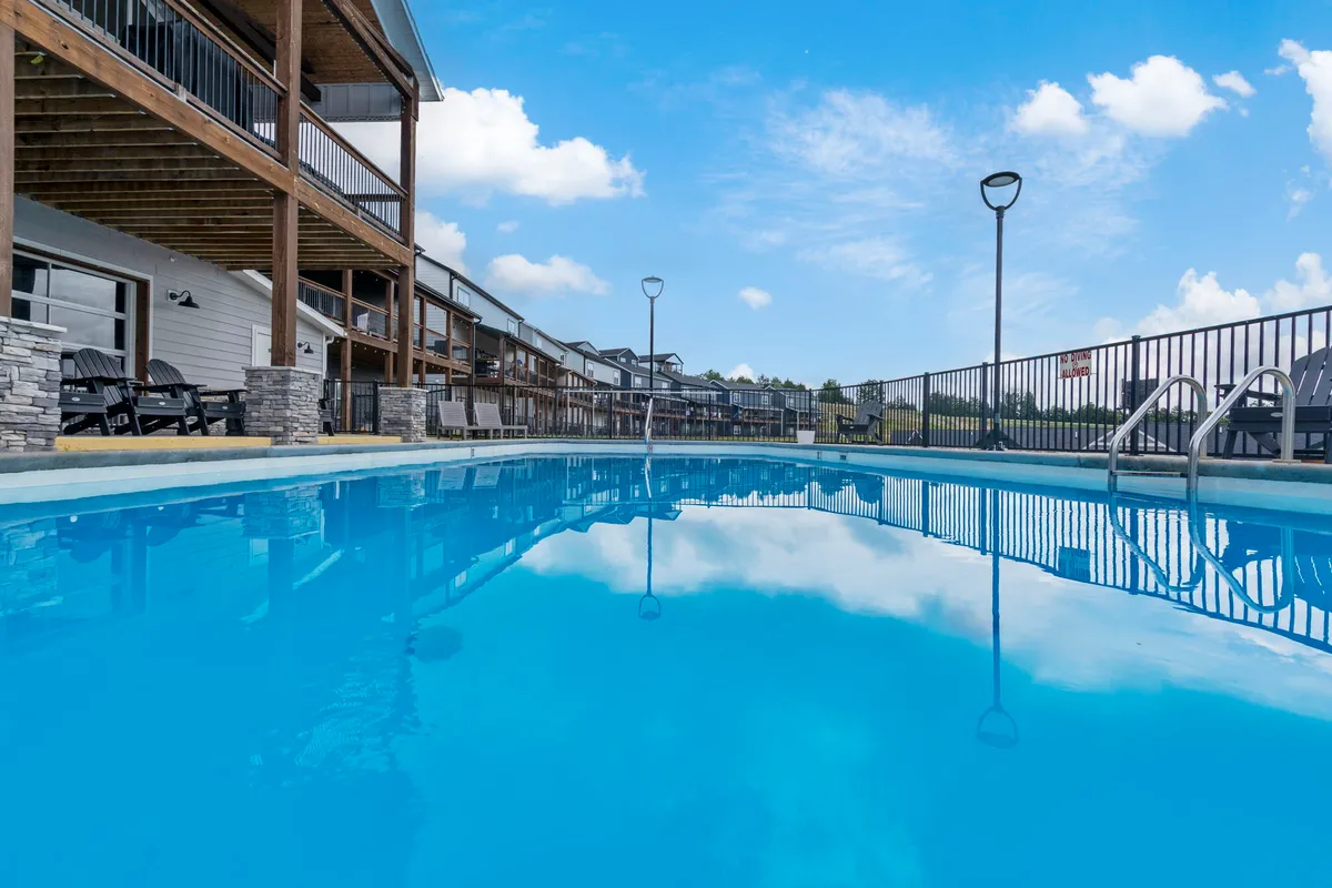 A sparkling outdoor swimming pool surrounded by a wooden deck and neighboring buildings, set against a clear blue sky. Lounge chairs are grouped around the pool for socializing and relaxation, with open spaces ideal for group gatherings.