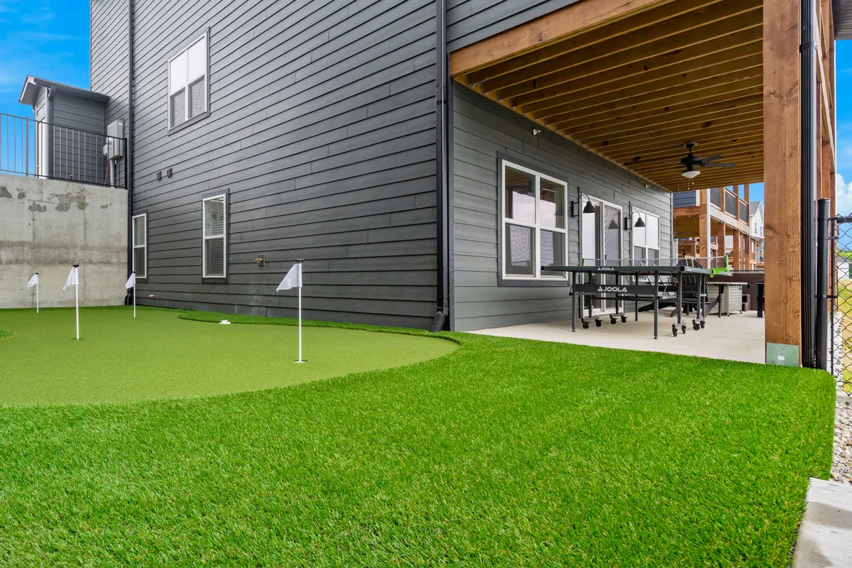 A sunlit outdoor space with a covered patio, dining table, and a green artificial putting green in the foreground. The setup invites both leisurely meals and fun putting competitions, capturing the spirit of resort-style amenities at home.