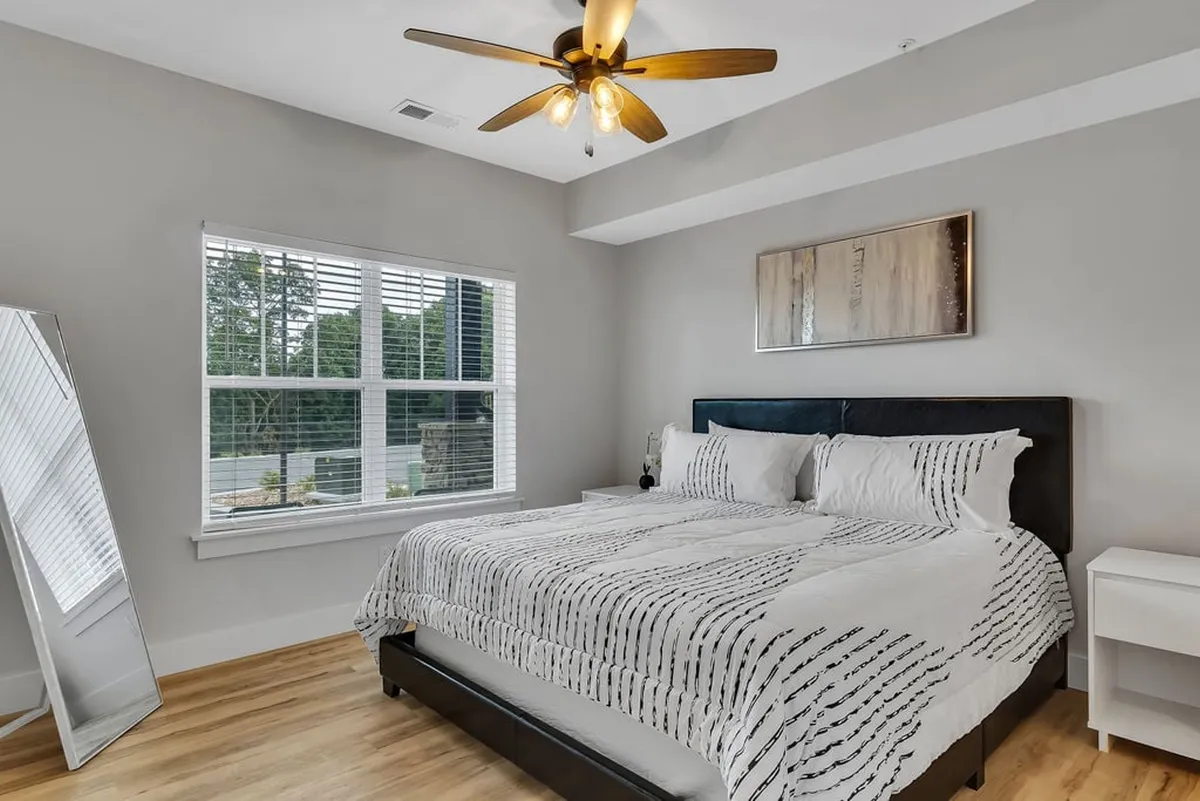 A serene king bedroom with a large bed covered in patterned bedding, a full-length mirror, a ceiling fan, and a large window streaming in natural light. The decor is peaceful and inviting, embodying the comfort and privacy promised to adults.