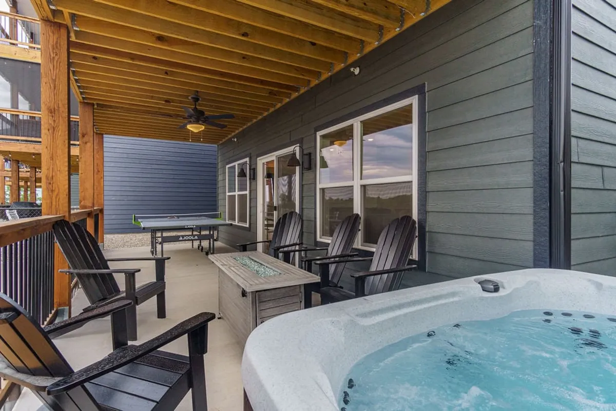 A lively outdoor patio scene featuring a bubbling hot tub, Adirondack chairs grouped for conversation, a ping pong table, and a ceiling fan overhead. The area is bathed in cheerful natural light, evoking a resort-like atmosphere perfect for unwinding and friendly games under the stars.